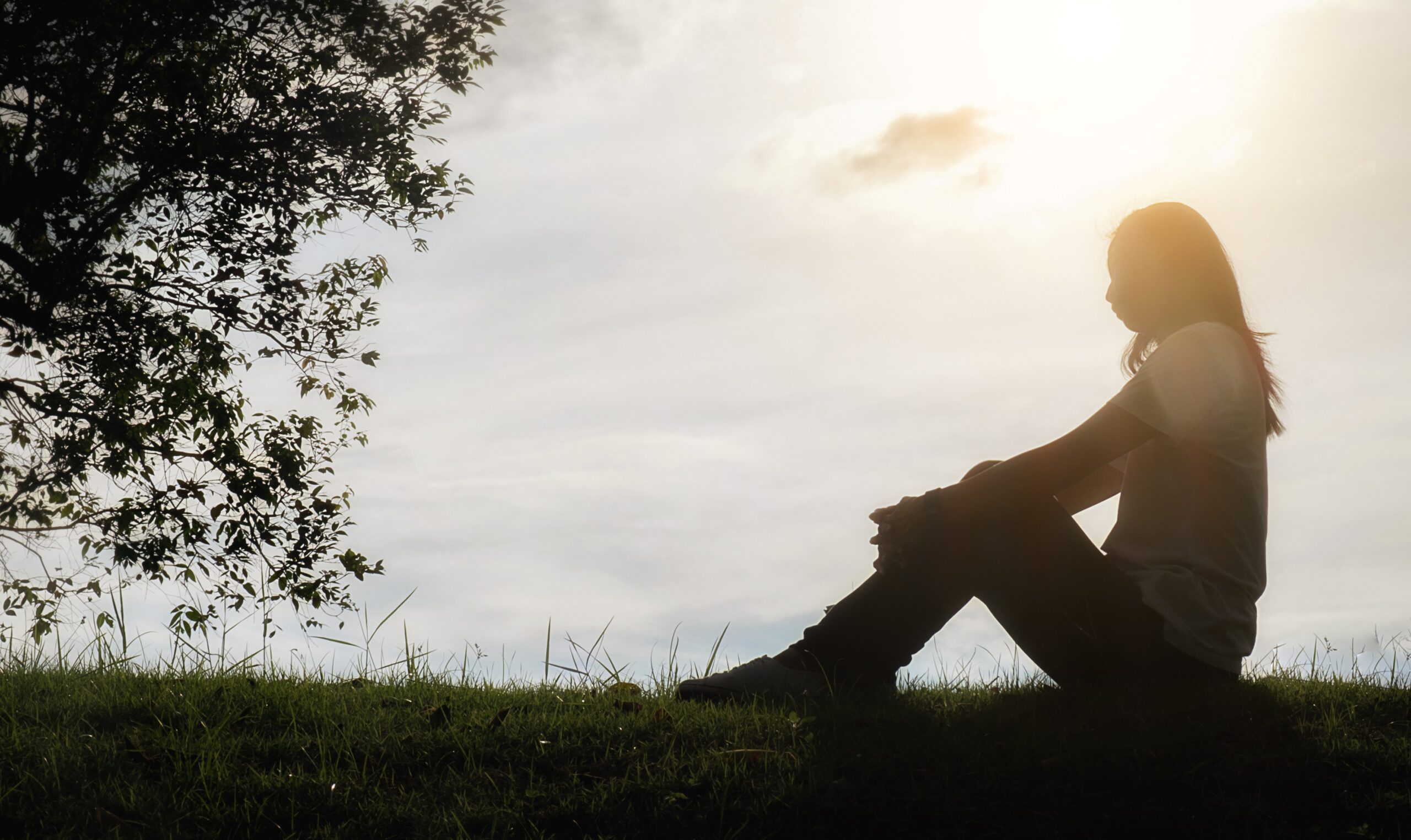 Sad and depressed woman sitting alone at the field during beautiful sunset with park background. Selective focus.